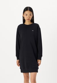Svart sweatshirt kjole laget av myk stoff, med lange ermer, rund halsutskjæring, og en liten brodert logo på brystet.