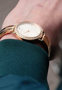 Gold-tone watch with a round dial, white face, and crystal accents. Attached to a thin, polished bracelet on a green sweater cuff.