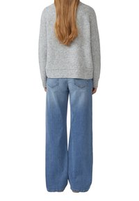 Vrouw met lang lichtbruin haar draagt een grijze textuurtrui en een broek met wijd uitlopende pijpen van blauwe denim, staande met haar rug naar de camera op een witte achtergrond.