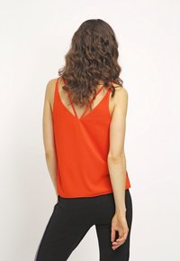 Top sin mangas rojo con un diseño de espalda en V y tirantes finos; hecho de una tela suave. Combinado con pantalones negros que tienen franjas laterales.