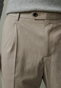 Nahaufnahme von beigen, maßgeschneiderten Hosen mit einer vorderen Falte, Gürtelschlaufen und Knopfverschluss, getragen mit einem hellgrauen Strickpullover.