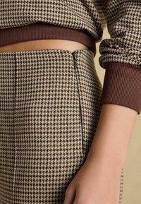 Brūns audums ar houndstooth rakstu un teksturētu virsmu, ar jostasvietu un rāvējslēdzēja detaļu kontrastējošā brūnā krāsā.