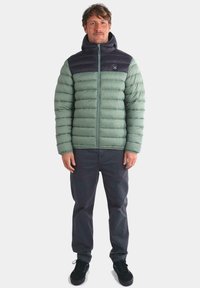 Chaqueta tipo plumón en verde claro con sección superior en verde oscuro, que cuenta con cierre de cremallera, capucha y textura acolchada. Llevada con pantalones oscuros y zapatos negros.