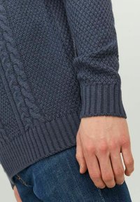 Marineblå strikket sweater med teksturerede mønstre, prydet med kabeldesign og ribbede cuffs. Nærbillede af ærmet med hånden hvilende på hoften.