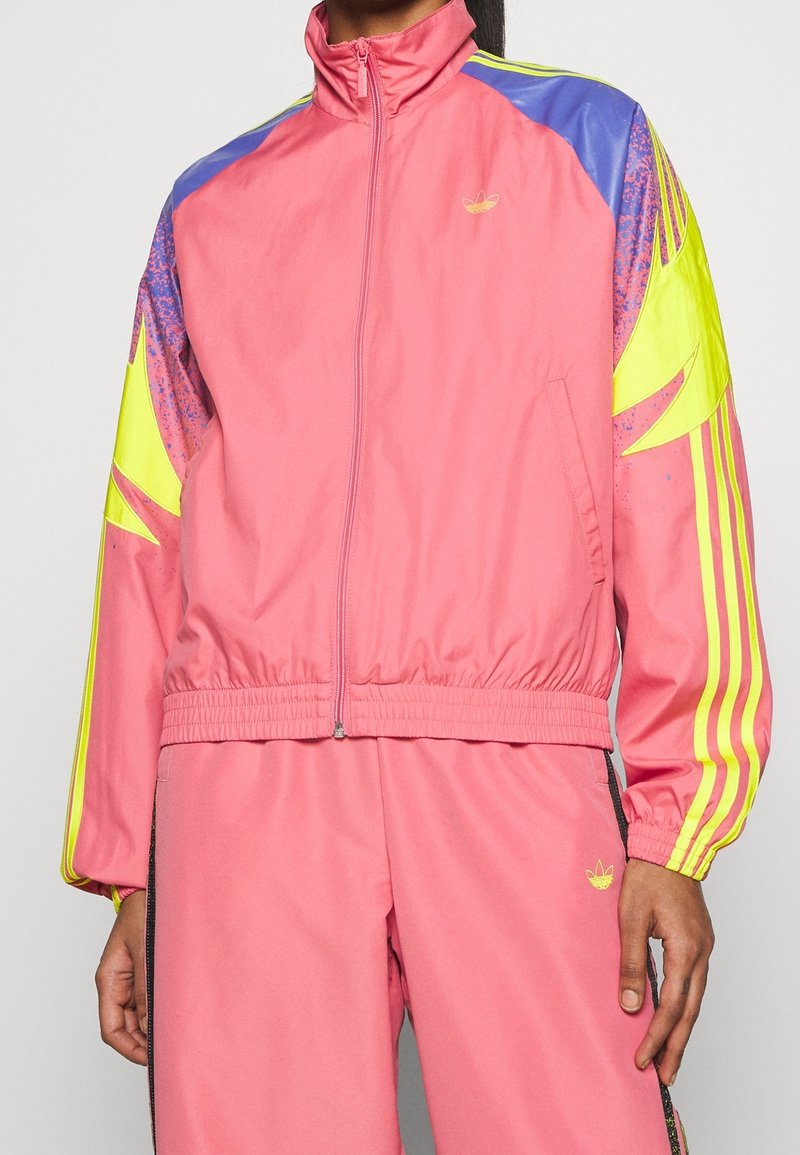 Pink zip-up jacket with yellow accents and blue shoulders. Features a fitted waist and vibrant graphic stripes on sleeves. Includes logo detail.