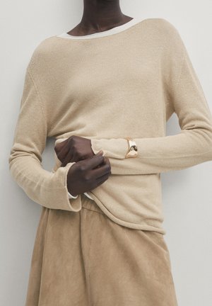 Dunkelhäutige Person trägt einen beigen Strickpullover und passende Wildlederhosen, zieht den Ärmel hoch; ein goldenes Manschettenarmband ist am Handgelenk sichtbar.