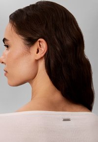 Femme aux longs cheveux foncés vue de profil et de dos, portant un haut blanc avec un petit logo Calvin Klein au niveau de l’encolure.