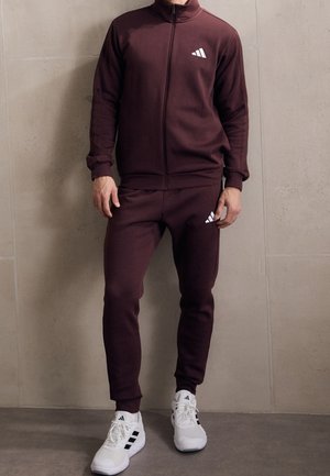 Uomo con tuta bordeaux con zip e scarpe da ginnastica bianche Adidas, in piedi contro un muro beige.