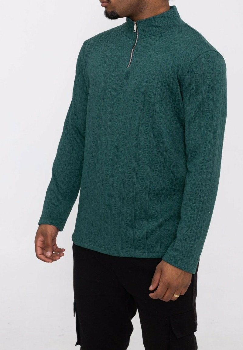 Pull en maille texturée verte avec un col montant, doté d'une courte fermeture éclair. Manches longues et design ajusté avec un motif en tricot torsadé.