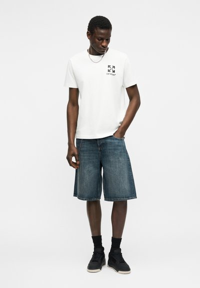 Άνδρας που φοράει λευκό t-shirt Off-White, τζιν σορτς μέχρι το γόνατο, μαύρα αθλητικά παπούτσια και μαύρες κάλτσες, στέκεται με το ένα χέρι στην τσέπη, κοιτώντας προς τα κάτω.