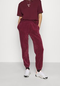 Pantalon de jogging en velours côtelé bordeaux avec taille et poignets élastiques, assorti à un tee-shirt de la même couleur. Des baskets blanches avec des accents néon complètent la tenue.