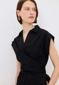 Blusa negra de manga corta con diseño cruzado y cuello, que presenta cierres de botones y una textura de tela suave.
