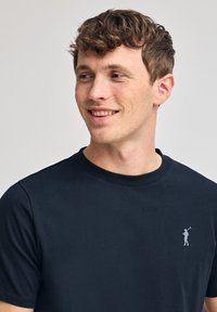 Mörkblå bomullströja med rund hals och korta ärmar. Har en liten golfergrafik på vänster bröst i ljusgrå.