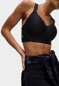 Sujetador deportivo negro con tela texturizada, detalle lateral recortado, tirantes ajustables y escote en V. Combinado con una chaqueta negra estampada atada a la cintura.