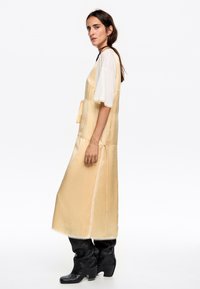 Abito giallo in satin con un dettaglio a fiocco, design senza maniche, spacco laterale, indossato sopra una t-shirt bianca, abbinato a stivali neri al ginocchio.