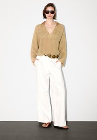 Maglione a coste beige, pantaloni bianchi a gamba larga, cintura spessa in oro e occhiali da sole scuri con infradito rosse. Tessuto liscio, vestibilità rilassata.