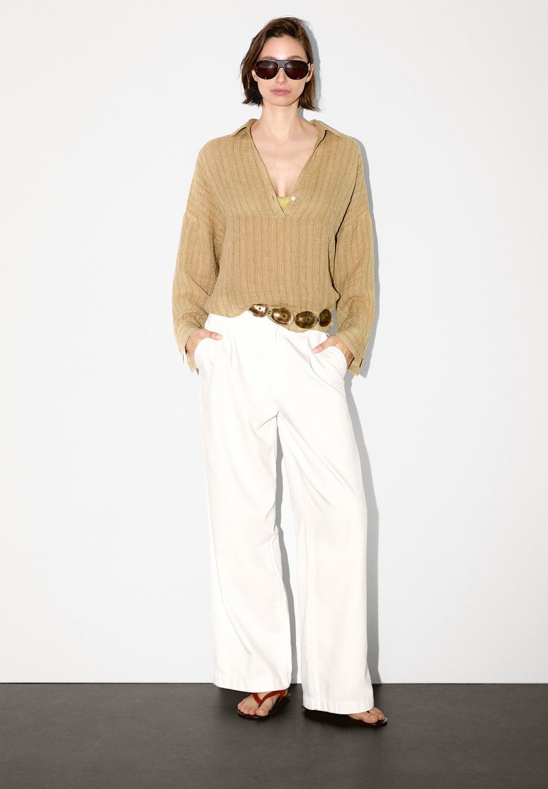 Maglione a coste beige, pantaloni bianchi a gamba larga, cintura spessa in oro e occhiali da sole scuri con infradito rosse. Tessuto liscio, vestibilità rilassata.