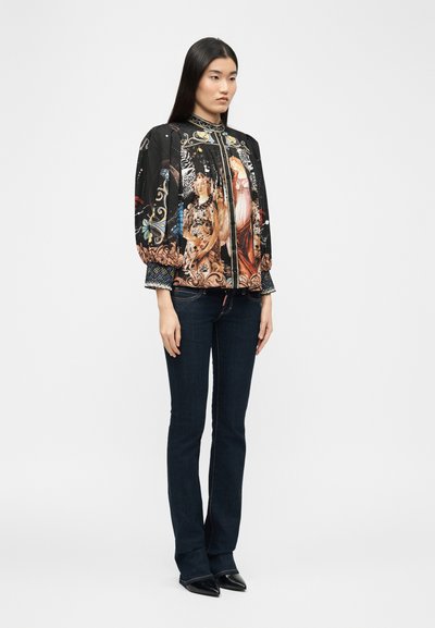 Femme portant une veste bomber noire avec des imprimés d'art de la Renaissance, un jean bleu foncé et des chaussures noires à bout pointu, debout devant un fond blanc uni.