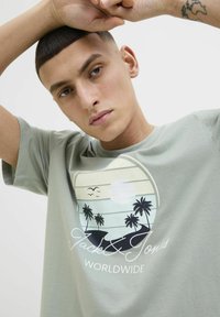 Camiseta de algodón verde claro con un gráfico de palmeras, un sol y el texto "Jack & Jones Worldwide". Mangas cortas y corte relajado.