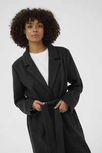 Černý kabát s páskem, šálovým límcem, dlouhými rukávy a bočními kapsami, předvedený přes bílý top. Hladká textura a přizpůsobený design.