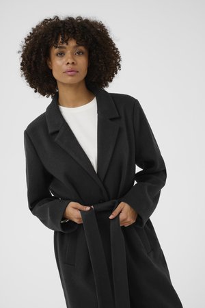 Schwarzer Mantel mit einem Schalkragen, langen Ärmeln und seitlichen Taschen, getragen über einer weißen Bluse. Glatte Textur und taillierte Passform.