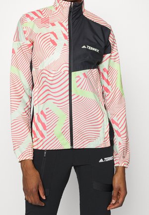 Person iført en farverig mønstret Adidas Terrex lynlåstrøje og sorte Adidas Terrex bukser, stående mod en ensfarvet baggrund.