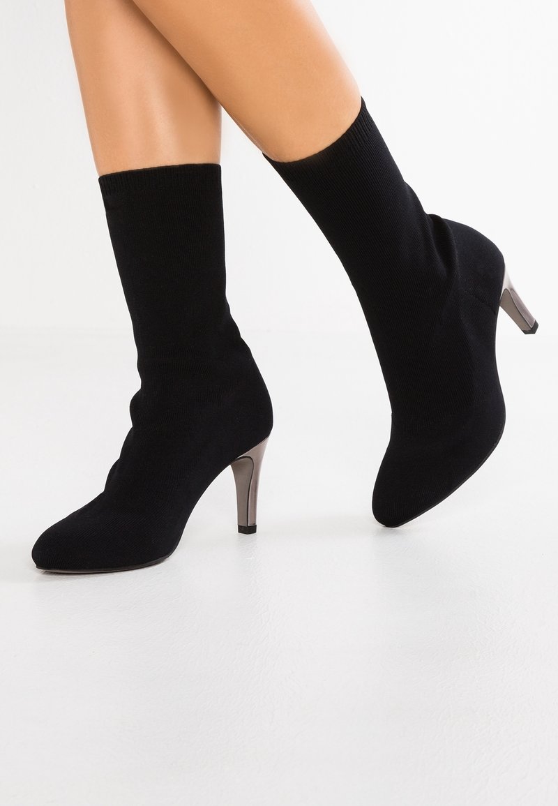 Bottes chaussettes en tissu noir avec talons argentés métalliques de hauteur moyenne portées aux pieds sur fond blanc.
