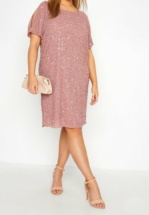 Femme portant une robe mauve à sequins mi-longue avec des manches à épaules dénudées, tenant une pochette beige et portant des escarpins beiges à brides autour de la cheville.