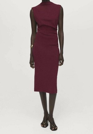 Femme portant une robe bordeaux sans manches, ajustée, longueur genou, avec des talons ouverts noirs et un bracelet doré au poignet gauche.