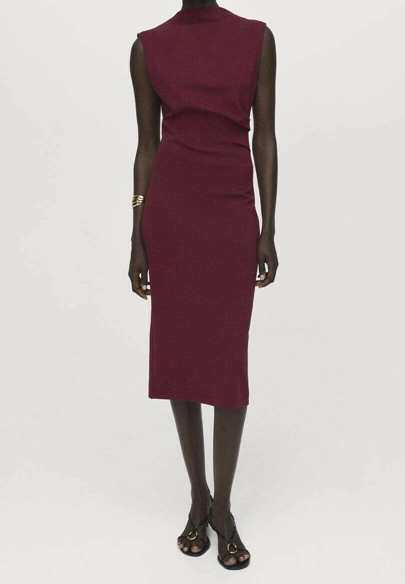 Femme portant une robe bordeaux sans manches, ajustée, longueur genou, avec des talons ouverts noirs et un bracelet doré au poignet gauche.