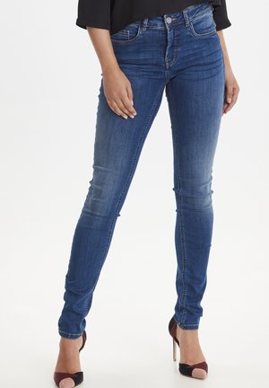 Jean en denim bleu mince avec une taille mi-haute, des détails de décoloration subtils et un design classique à cinq poches, associés à des chaussures à talons hauts.