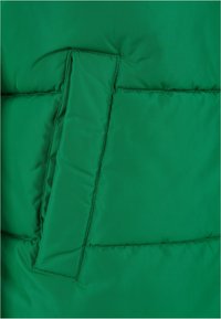 Grön puffjacka med texturerad, slät yta. Har en vikt ficklock, synliga sömmar och en fluffig quiltad design.