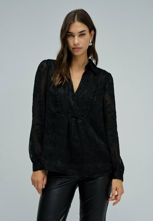 Blouse noire transparente avec broderies florales, collerette et manches longues. Associée à un pantalon en cuir noir ajusté.