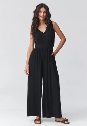 Kobieta o długich kręconych włosach, ubrana w bezrękawnikowy czarny kombinezon z szerokimi nogawkami i brązowymi sandałami, stojąca z jedną ręką w kieszeni.