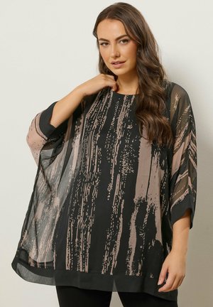Frau trägt eine lockere, transparente schwarze Tunika mit abstrakten beigen Pinselstrichmustern und steht vor einem einfarbigen hellen Hintergrund.