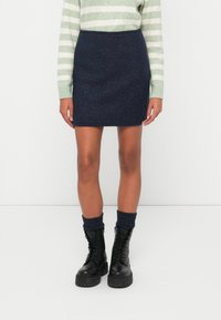 Minifalda texturizada azul marino con un corte ajustado. Combinada con botas negras hasta el tobillo con cordones y calcetines de canalé azul marino. Suéter a rayas verdes y blancas.