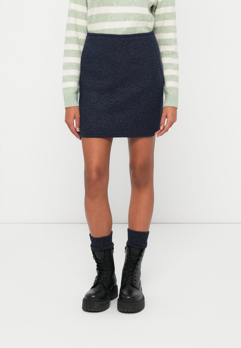 Minifalda texturizada azul marino con un corte ajustado. Combinada con botas negras hasta el tobillo con cordones y calcetines de canalé azul marino. Suéter a rayas verdes y blancas.