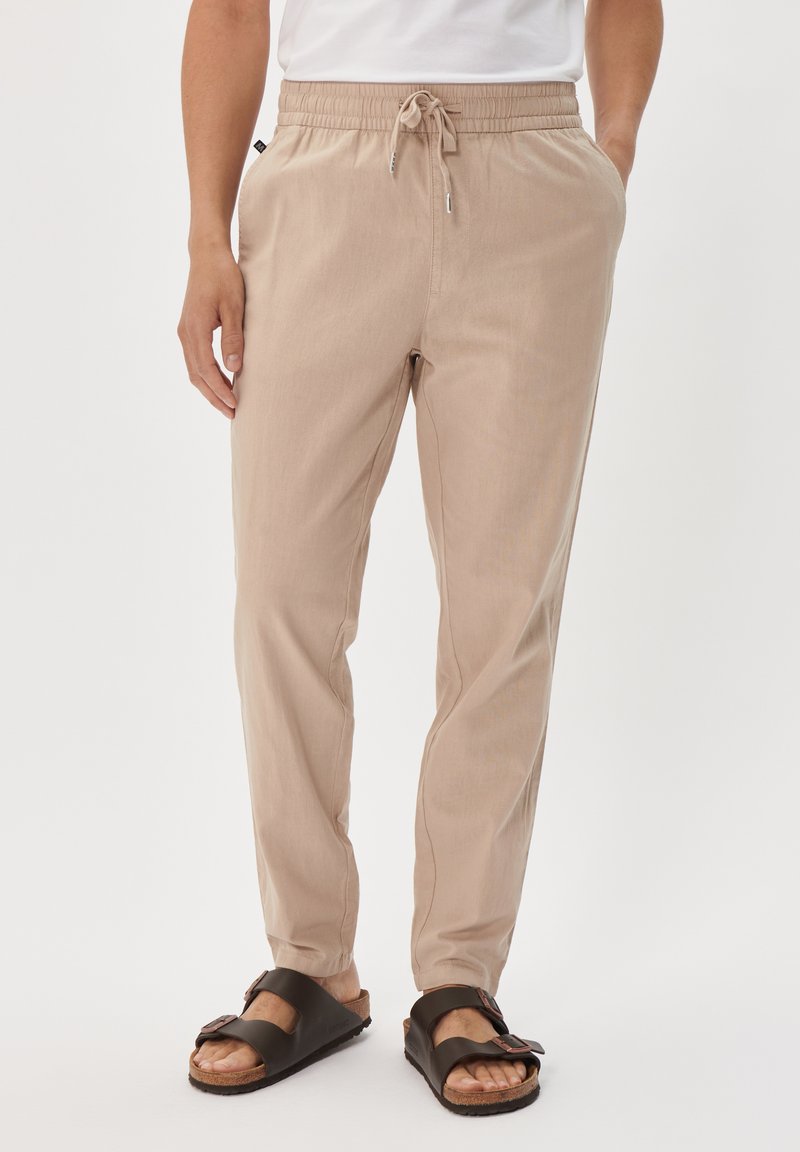 Beige, lightweight pants with an elastic waistband and drawstring. Smooth texture, tapered legs, side pockets. Paired with brown sandals.