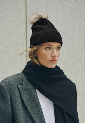 Junge Frau mit schwarzer Strickmütze mit Bommel, dunklem Schal und grauem Mantel, blickt nachdenklich zur Seite vor hellem Hintergrund.