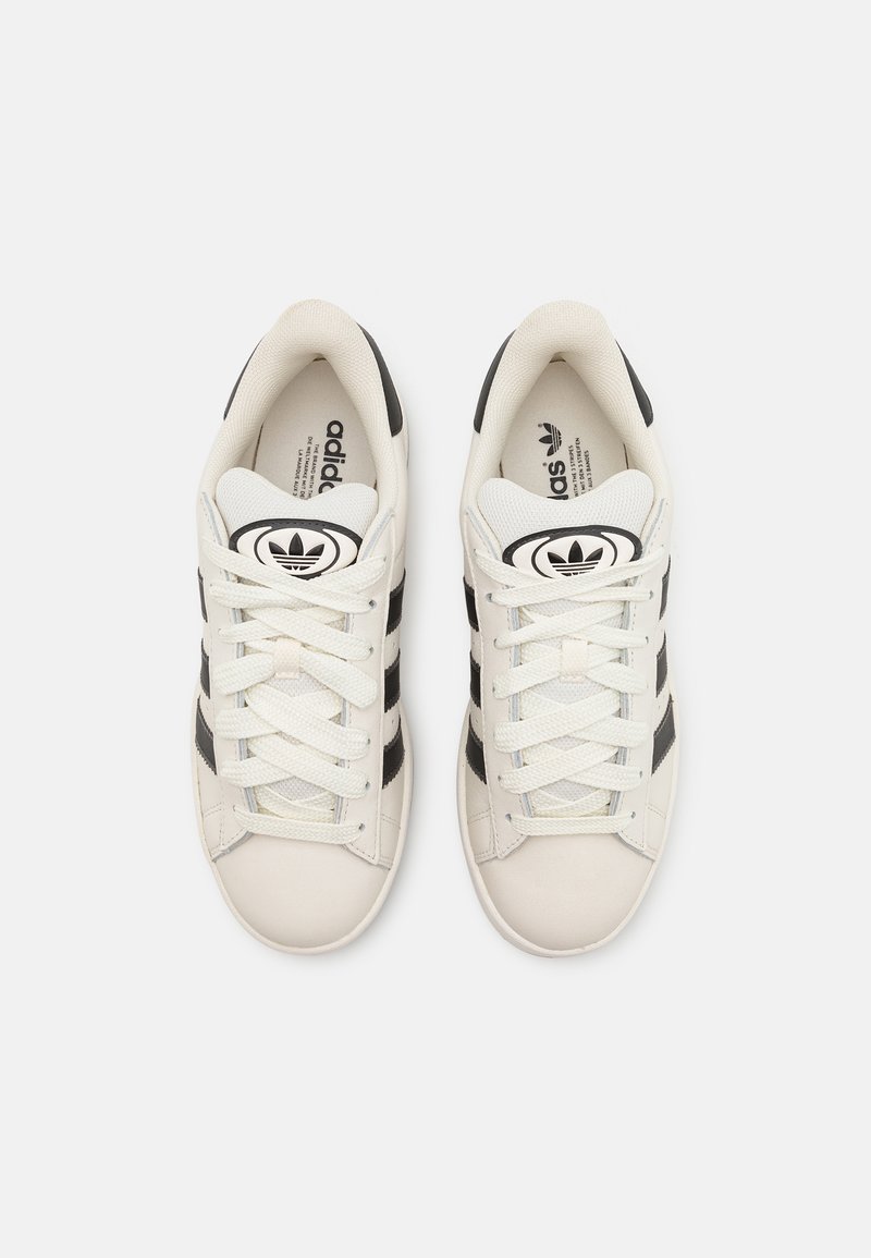 Baskets de sport en cuir blanc avec des rayures noires, présentant un bout rond, des lacets texturés et un logo sur la languette et la semelle intérieure.