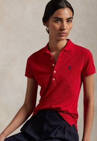Red polo shirt made of textured cotton, featuring a button placket and blue embroidered logo on the left chest. Pairing with dark pants.