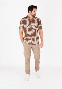Camicia a maniche corte, con motivo marrone e crema, abbinata a pantaloni cargo color kaki e sneaker bianche. Presenta una chiusura con bottoni e una vestibilità comoda.