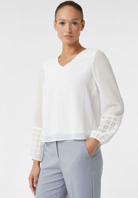 Biała bluzka z dekoltem w kształcie litery V i przezroczystymi rękawami, z teksturowanym wzorem kratki na mankietach. W połączeniu z jasnoszarymi spodniami.