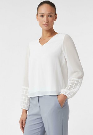 Weiße Bluse mit V-Ausschnitt und transparenten Ärmeln, mit einem strukturierten Gittermuster an den Manschetten. Kombiniert mit hellgrauen Hosen.