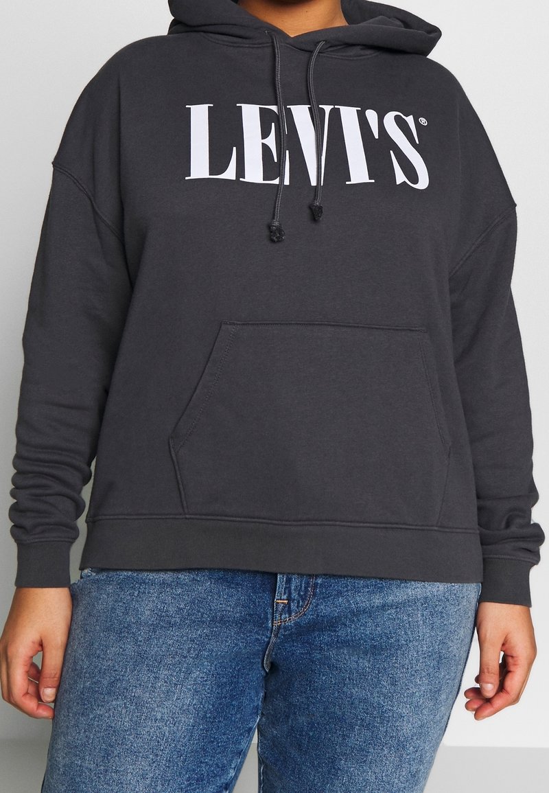 Personne portant un sweat à capuche noir Levi's avec une poche avant et un jean bleu, debout devant un fond clair uni.