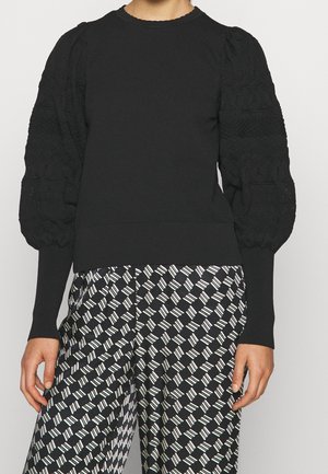 Kvinde iført en sort tekstureret sweater med puf-ærmer kombineret med sorte og hvide mønstrede vidde bukser.