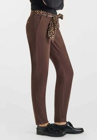 Pantaloni marroni con una vestibilità slim, polsini elastici e una sciarpa con stampa leopardata annodata in vita, abbinati a scarpe nere.