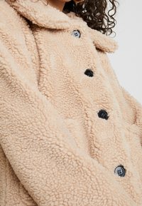 Detailní záběr na osobu oblečenou v béžovém, chlupatém kabátu s límcem, který má černé knoflíky a texturovanou látku.