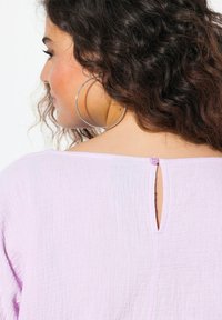 Blusa lavanda caratterizzata da un design tessuto testurizzato con uno scollo a chiave sul retro e un piccolo dettaglio con bottone abbinato. Orecchini a cerchio argentati visibili.