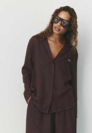 Woman with curly hair wearing large dark sunglasses and a loose dark brown button-up shirt with matching pants, standing against a white background.
